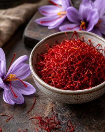 Crvene niti šafrana u keramičkoj činiji pored ljubičastih cvetova Crocus sativus na tamnoj rustičnoj podlozi.