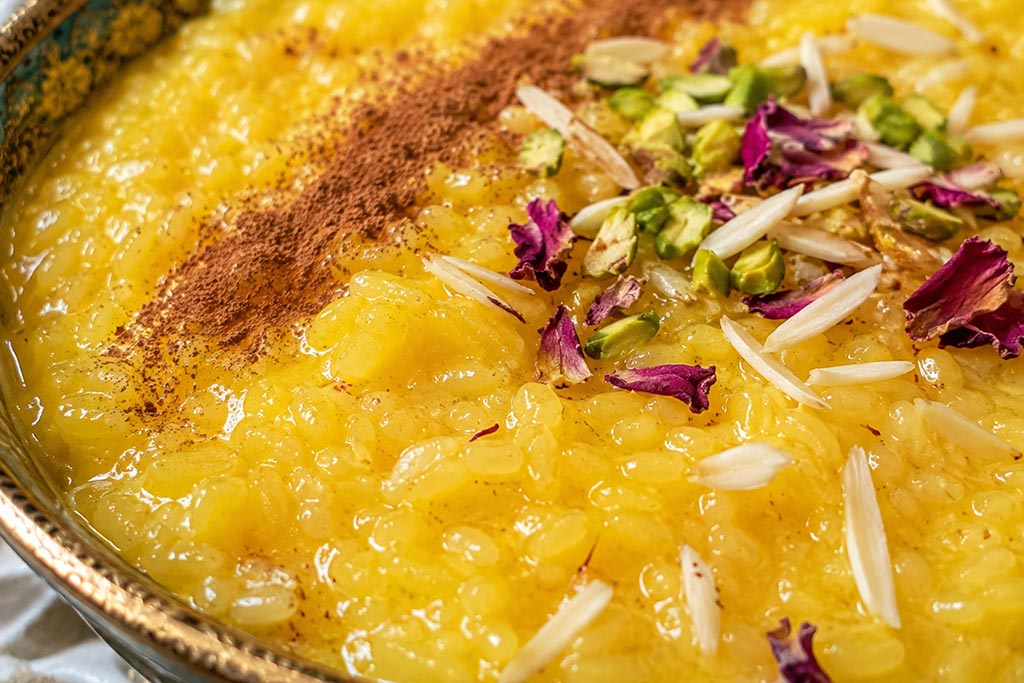 Krupni kadar kremaste teksture Sholeh Zarda, iranskog pudinga od pirinča sa šafranom, ukrašenog cimetom, pistaćima, bademima i suvim laticama ruže.