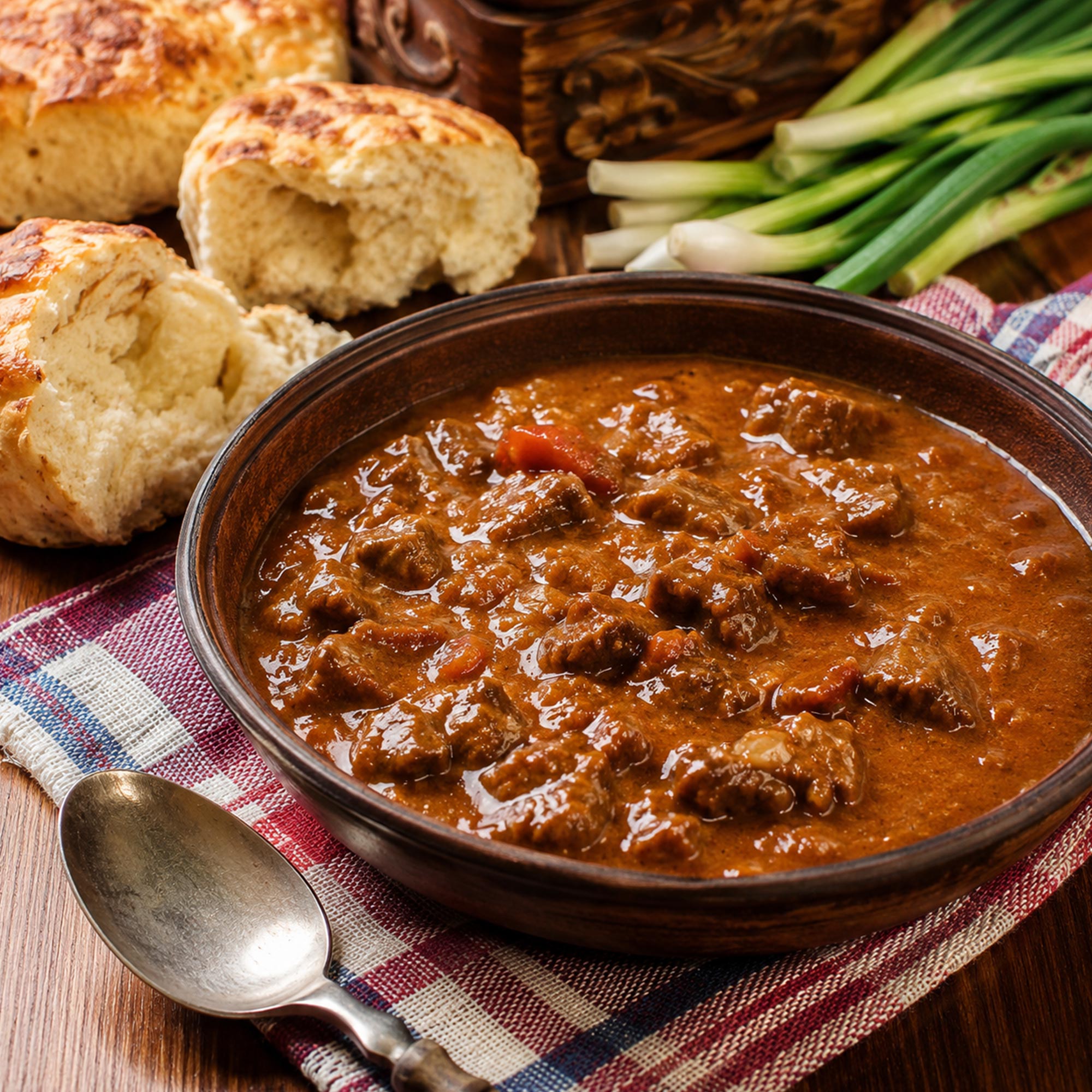 Gulaš poslužen u tradicionalnom tanjiru uz domaću pogaču, mladi luk i kariranu krpu na rustičnom drvenom stolu.
